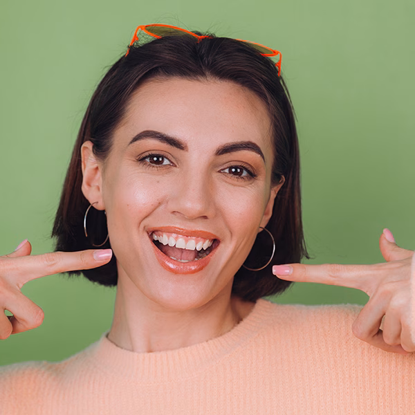 Young stylish woman in casual peach sweater and orange glasses isolated on green olive background positive smiling pointing on white teeth with index fingers copy space